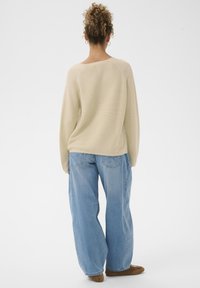 Personne avec des cheveux bouclés portant un haut à manches longues beige, un jean bleu clair ample et des chaussures marron, se tenant de dos sur un fond blanc.