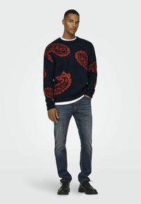 Marineblauwe trui met rode bloemenpatronen, ronde hals, gebreide textuur. Gecombineerd met vergrijsde blauwe jeans en zwarte veterschoenen.