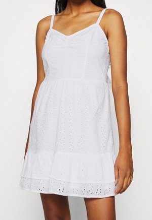 Kobieta w białej, bezrękawnikowej sukience z ażurowym wzorem i falbanką na dole, stojąca ze swobodnie opuszczonymi ramionami na tle jednolitego tła.