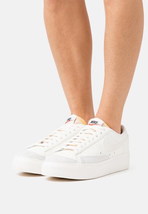 Baskets blanches avec un bout en daim texturé, un corps en cuir lisse et une semelle en caoutchouc épaisse ; elles présentent des accents rouges et bleus sur la languette.