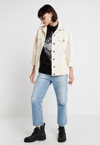 Lys beige denimjakke med knaplukning, båret over en sort grafisk t-shirt. Mellemblå slidte jeans og sorte snørebåndsstøvler fuldender outfittet.