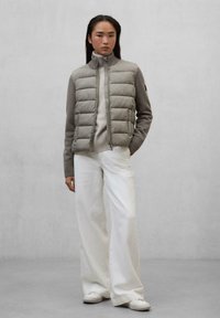 Gilet imbottito grigio chiaro con maniche a costine, chiusura con zip e colletto alto, abbinato a pantaloni bianchi a gamba larga e scarpe da tennis bianche.