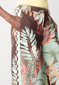 Floral trousers in brown featuring green, red, and beige plant patterns, a checkered side detail, and a side pocket. Made from lightweight fabric.