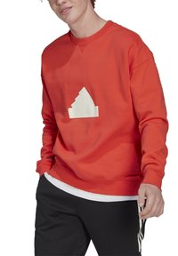 En ung person som bär en röd sweatshirt med en vit geometrisk logotyp, svarta byxor med vita ränder och en vit skjorta som tittar fram.
