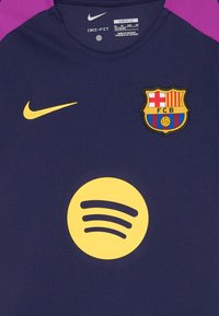 Nike Dri-FIT dres v tmavomodrej farbe, s fialovými akcentami na ramenách, logom Barcelony, žltým logom Spotify v tvare kruhu a elegantnou, štruktúrovanou tkaninou.