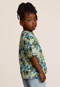 Chemise à manches courtes avec une base verte, ornée d'un motif floral bleu, jaune et vert. Tissu doux, coupe ample, col rond.