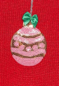 Pull rouge décoré d'un ornement en sequins roses et dorés avec un nœud vert au-dessus, affichant des motifs circulaires et des détails texturés.