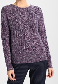 Maglione a maglia viola con un motivo texturizzato e polsini a coste, con scollatura rotonda. Abbinato a pantaloni a righe.