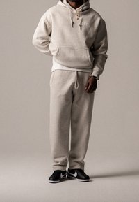 Mężczyzna w jasnoszarej bluzie z kapturem i dresowych spodniach, w czarno-białych trampkach Nike, stoi na tle bez wzorów, z jedną ręką schowaną za plecami.