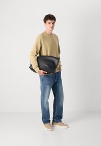 Zwarte leren schoudertas vastgehouden door een persoon in een beige sweatshirt en blauwe jeans. Gevoelstruur leren met een eenvoudig, gestroomlijnd ontwerp.