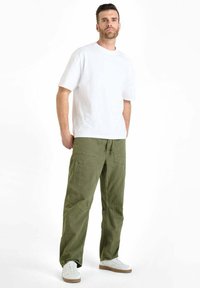 Man staat met handen in de zakken, draagt een witte kortekeer shirt, olijfgroene cargobroek en witte sneakers tegen een witte achtergrond.