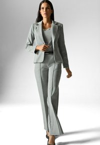 Vrouw loopt zelfverzekerd in een op maat gemaakt lichtgrijs pak met een blazer, bijpassende broek, riem en zwarte hakken op een effen achtergrond.