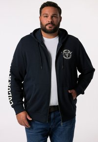 Hoodie zippé bleu marine avec capuche à cordon, poches avant et logo rond sur la poitrine ; présente du texte blanc le long de la manche.