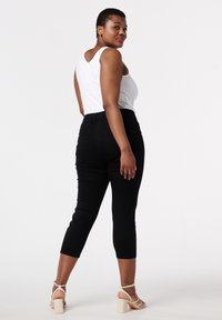 Pantalons capri noirs ajustés avec une taille élastique, associés à un débardeur blanc et des sandales à talons blocs beiges, exposés de dos.
