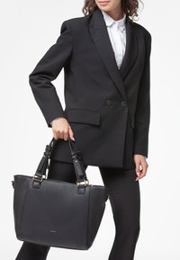 Borsa tote nera in pelle con superficie testurizzata, dettagli in metallo dorato, manici piatti e forma rettangolare, tenuta da una persona in abbigliamento business.