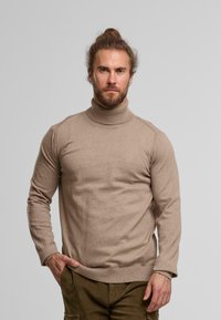 Mann mit Bart und zurückgebundenem Haar, der einen beige Rollkragenpullover und braune Hosen trägt, steht vor einem einfarbigen grauen Hintergrund.