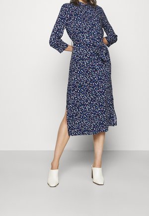 Frau trägt ein marineblaues Midikleid mit mehrfarbigem Punktmuster, dreiviertel Ärmeln, seitlichem Schlitz, gebundenem Taillengürtel und weißen Mules mit Absatz.