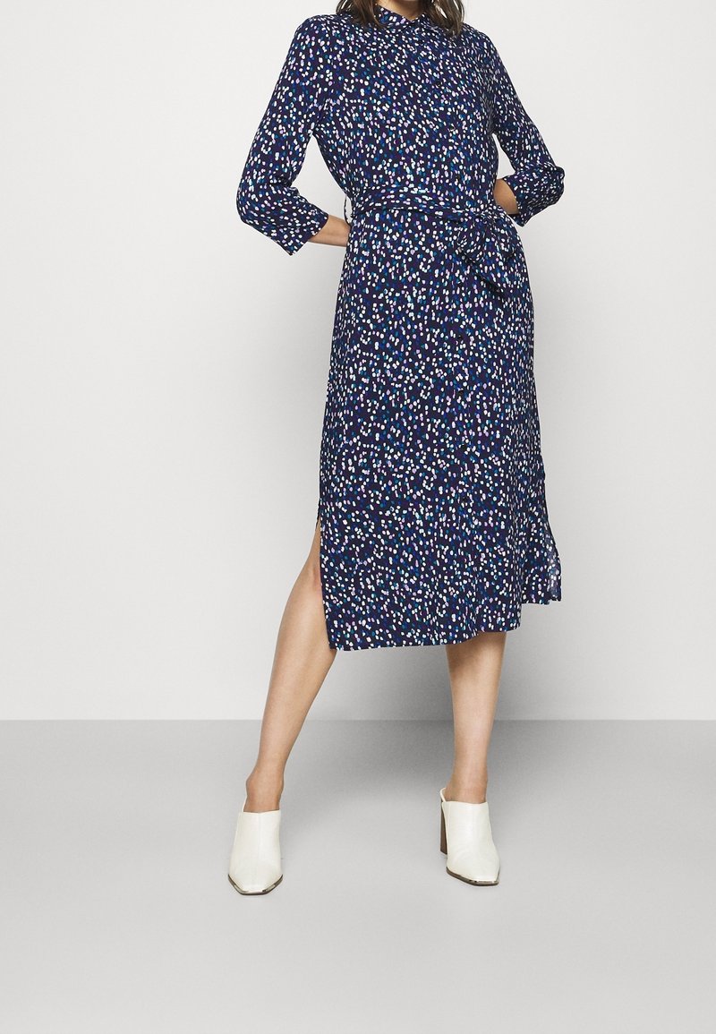 Femme portant une robe midi bleu marine avec un motif à pois multicolore, manches trois-quarts, fente latérale, taille nouée, et mules blanches à talons.