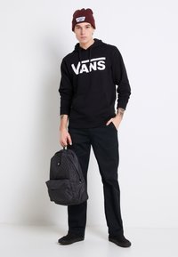 Felpa nera con grande logo bianco "VANS", pantaloni neri e sneaker scure, mentre tiene uno zaino nero texturizzato con tasca frontale. Berretto bordeaux.