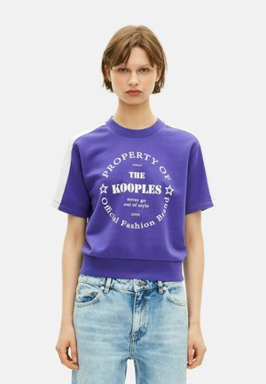 Jeune personne portant un sweatshirt violet à manches courtes avec du texte blanc et un jean bleu clair, se tenant devant un fond blanc uni.