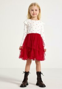 Rotes Tüllkleid mit einem weißen Langarmoberteil, das mit roten Herzmustern verziert ist. Stylisch kombiniert mit braunen Schnürstiefeln. Rüschendetails an den Ärmeln.