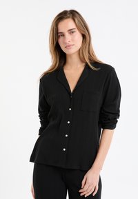 Czarna koszula z guzikami, o fakturowanym prążkowanym wzorze, z kołnierzykiem typu złożonego, długimi rękawami i kieszonką na piersi. Białe guziki jako akcenty.