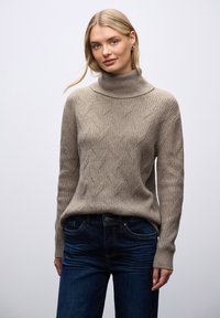 Beżowy sweter robiony na drutach z wysokim kołnierzem, ozdobiony fakturowanym wzorem w diamenty, połączony z ciemnoniebieskimi dżinsami.