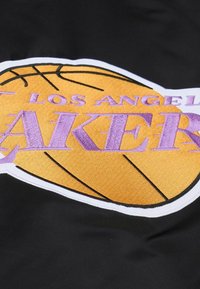Schwarzer Stoff mit einem auffälligen gestickten Logo der Los Angeles Lakers, das einen goldenen Basketball und lila Text zeigt. Strukturierte Oberfläche mit kontrastierenden Farben.