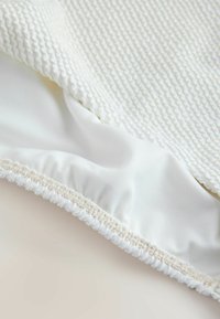 Balta tekstūruota medžiaga su briaunuotu paviršiumi ir lygiu apačia. Kraštai turi surinktą elastinę juostelę, užtikrinančią patikimą prigludimą. Minkšta, neutrali spalva.