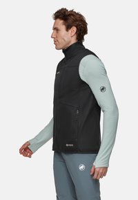 Schwarzer Softshell-Weste mit hohem Kragen, einem Reißverschlussfach vorne und zwei Seiten Taschen. Getragen über einem hellblauen, gerippten Langarmshirt.