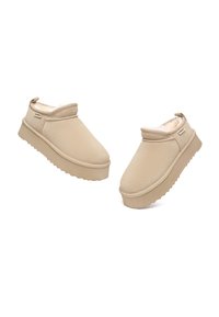 Beige Slippers mit einer strukturierten Oberseite, weicher Fütterung und einer dicken, gerippten Sohle. Besitzt ein kleines Logo an der Seite und ein umgeschlagenes Schafdesign.