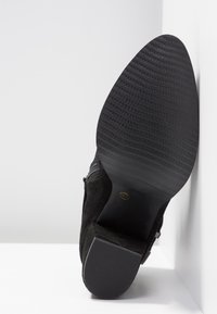 Botte en daim noir montrant une semelle texturée et un petit talon carré, inclinée sur fond blanc d'angle.