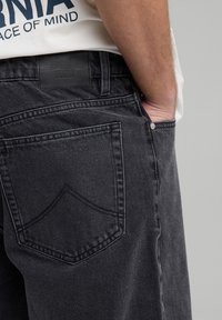 Lähikuva henkilöstä, joka käyttää mustia farkkuja ja on laittanut yhden käden etutaskuun. Hänellä on päällään valkoinen paita, joka on osittain laitettu sisään.