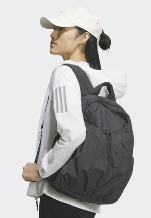 Zaino in nylon nero con chiusura a coulisse regolabile, spallacci imbottiti e forma elegante e arrotondata. La modella indossa una giacca leggera e un berretto.