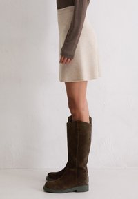 Person trägt einen beigen Rock, ein braunes Langarmshirt und hohe dunkelbraune Wildlederstiefel und steht vor einer schlichten weißen Wand.