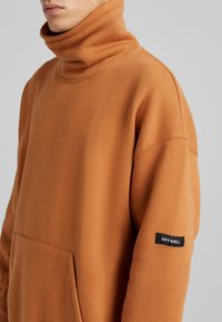 Sudadera de gran tamaño en color naranja quemado, fabricada en tejido suave, con un alto cuello, bolsillo lateral y etiqueta negra "APPAREL" en la manga.