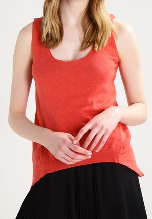 Femme portant un haut en maille rouge sans manches et une jupe plissée noire, les mains doucement jointes à la taille.