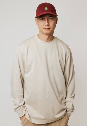 Młody mężczyzna z rękami w kieszeniach, ubrany w kremową koszulę z długim rękawem i czerwoną czapkę z zielonym emblematem drzewa, stojący na jednolitym tle.