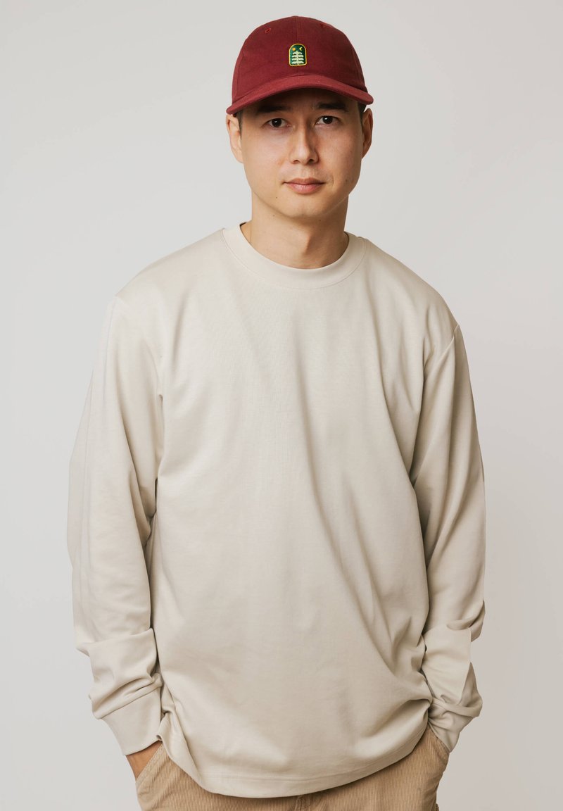 Młody mężczyzna z rękami w kieszeniach, ubrany w kremową koszulę z długim rękawem i czerwoną czapkę z zielonym emblematem drzewa, stojący na jednolitym tle.