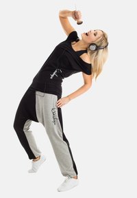 Black off-shoulder top and gray-black color-block joggers adorned with script. Model wears headphones and white sneakers, holding a microphone.