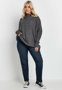 Maglione grigio in grandi dimensioni a trama cable-knit con scollatura alta, abbinato a jeans a gamba dritta blu scuro e sneaker nere. Presenta polsini a costine.