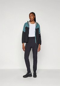 Črno in modrozelena vetrnica z zadrgo, kombinirana s črnimi oprijetimi hlačami in črnimi supergami. Jakna ima sijočo srebrno notranjo podlogo.