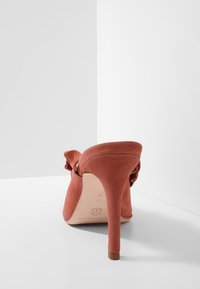 Rustfarbenes Wildleder-Pump mit Absatz und dekorativem Schleifen, von hinten vor einem weißen Hintergrund betrachtet.