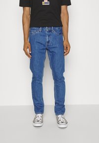 Blå denimjeans med slim fit, med fem fickor och kontrasterande sömmar, som bärs med mönstrade sneakers och en svart t-shirt.