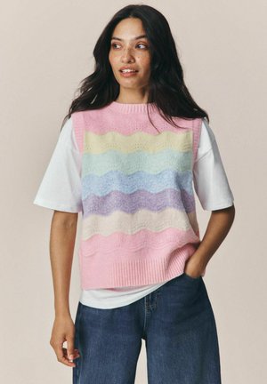 Kvinde med langt mørkt hår iført en pastelstribet strikket vest over en hvid t-shirt og blå jeans, stående med den ene hånd i lommen.