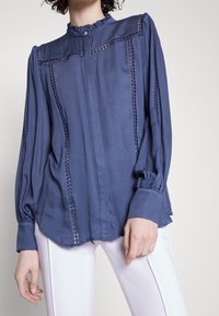 Femme portant un chemisier bleu à manches longues avec des empiècements en dentelle et des poignets froncés, associé à un pantalon blanc sur un fond uni.