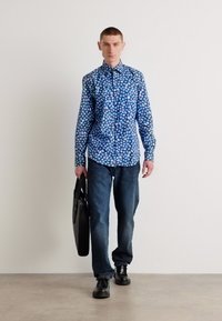 Chemise bleu à manches longues avec un motif floral, associée à un jean en denim noir. Le mannequin porte un sac noir et des chaussures noires.