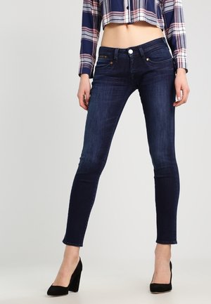 Femme portant un jean skinny bleu foncé, des escarpins noirs et une chemise à carreaux courte à manches longues, debout sur un fond uni.