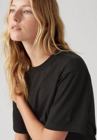 Tricou negru cu mânecă scurtă, cu guler rotund, dintr-un material moale, logo subtil pe mânecă și o croială lejeră, prezentat cu părul desfăcut și un fundal neutru.