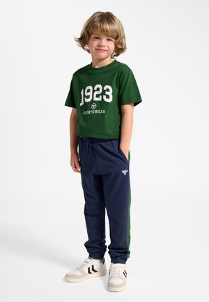 Młody chłopiec o blond włosach, ubrany w zieloną koszulkę z napisem "1923 Sportswear", granatowe i zielone spodnie dresowe oraz białe trampki, stoi z rękami w kieszeniach.